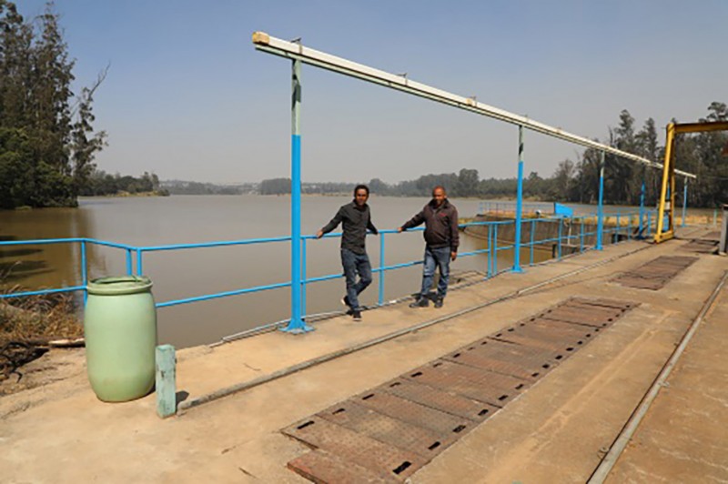 Two Hub members stand against the blue railings on the edge of the dam, with the expanse of the river behind